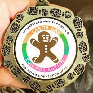 Photo of a medal for Gingerbread Man Running Company's Run for Autism Connection Labor Day Race September 5, 2022. Brushed metallic circle with shoe prints framing a white center that has a smiling Gingerbread Man in mid sprint with arms in the air and feet off the ground.