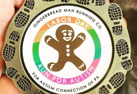 Photo of a medal for Gingerbread Man Running Company's Run for Autism Connection Labor Day Race September 5, 2022. Brushed metallic circle with shoe prints framing a white center that has a smiling Gingerbread Man in mid sprint with arms in the air and feet off the ground.