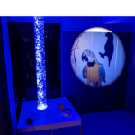 Photo of sensory room with dim blue lights and images of parrots projected on wall next to a bubble tube machine
